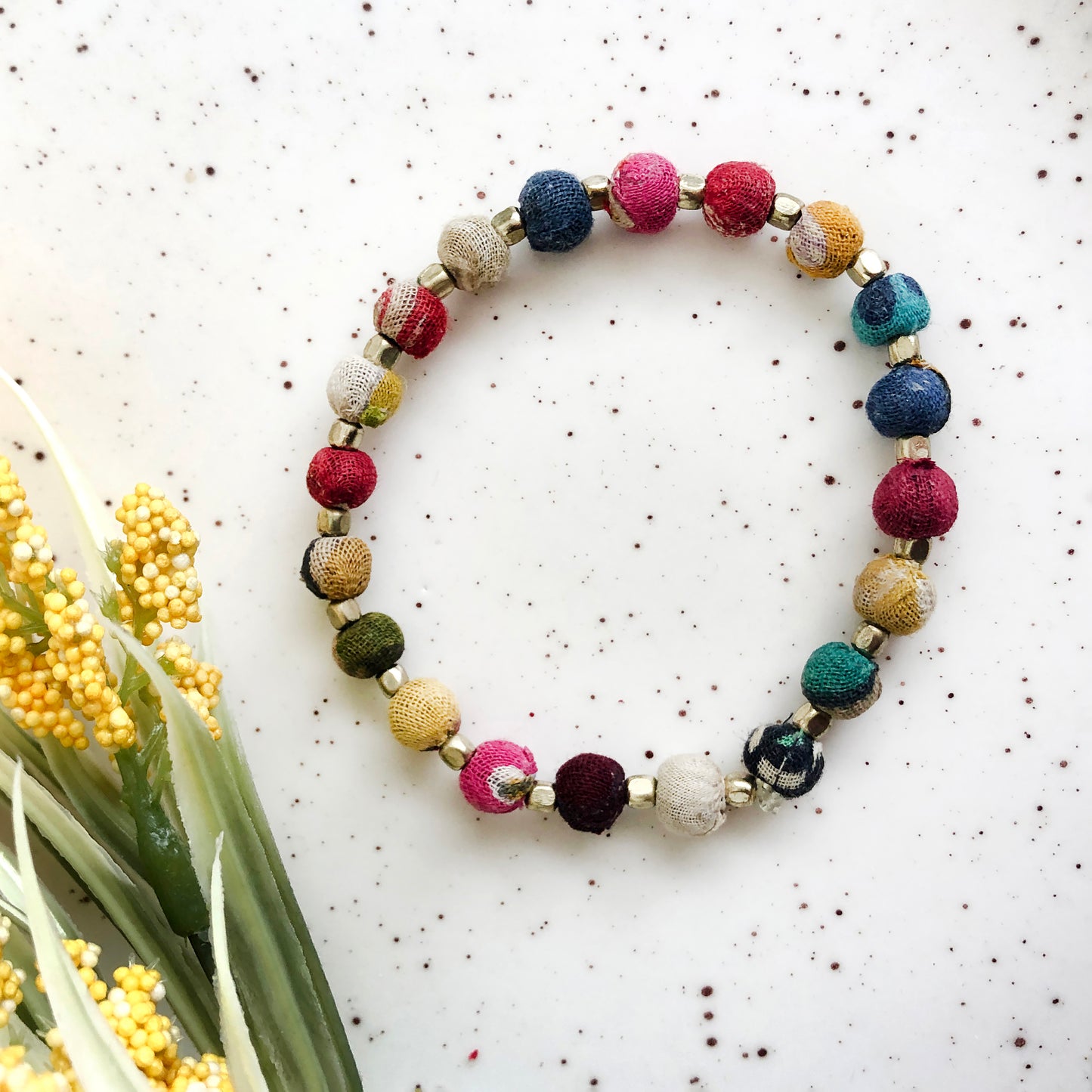 Dotted Kantha BraceletA bracelet made from small colorful textile-wrapped beads is shown next to yellow flowers.