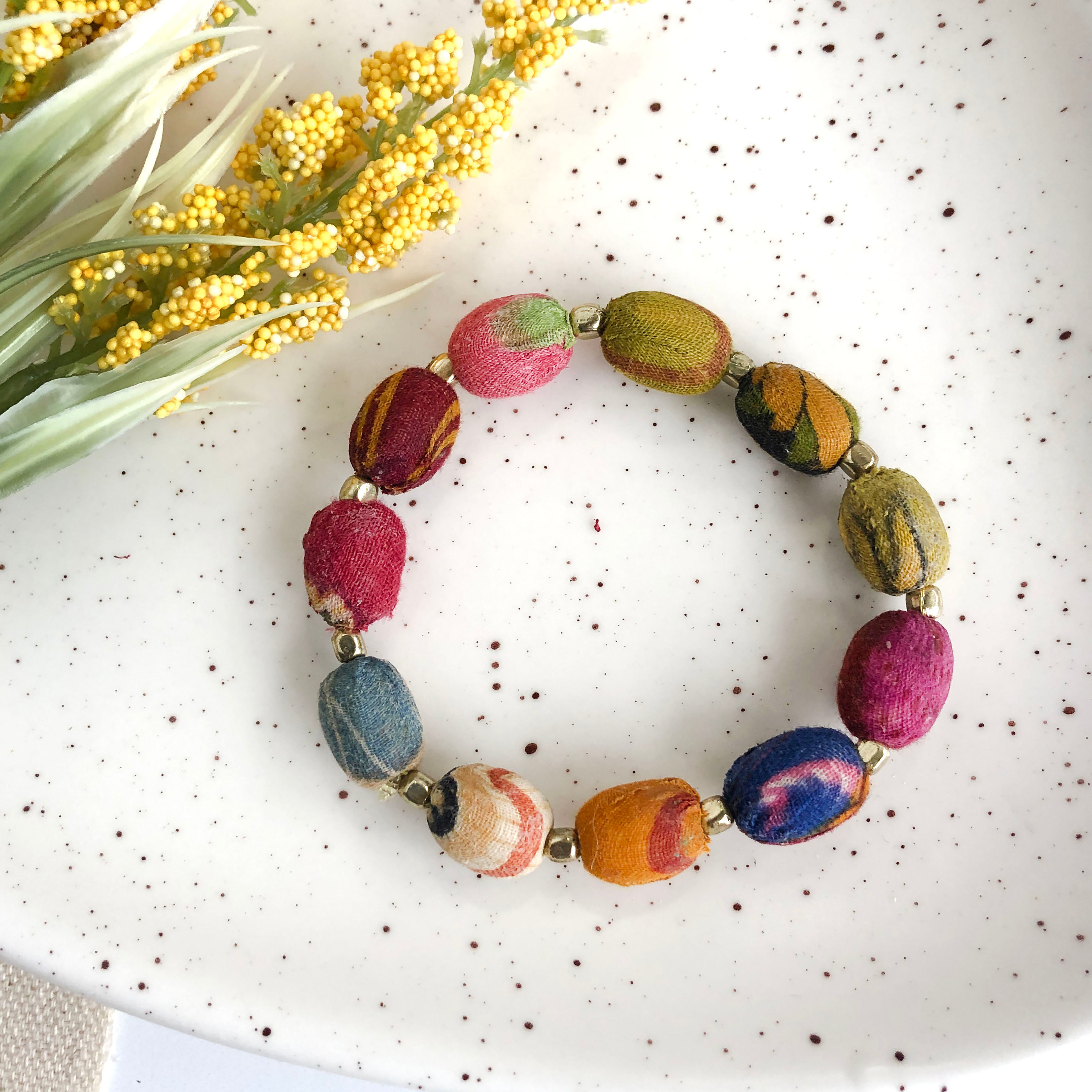 A colorful strand of textile-wrapped oblong beads alternates with small gold beads to create a bracelet.