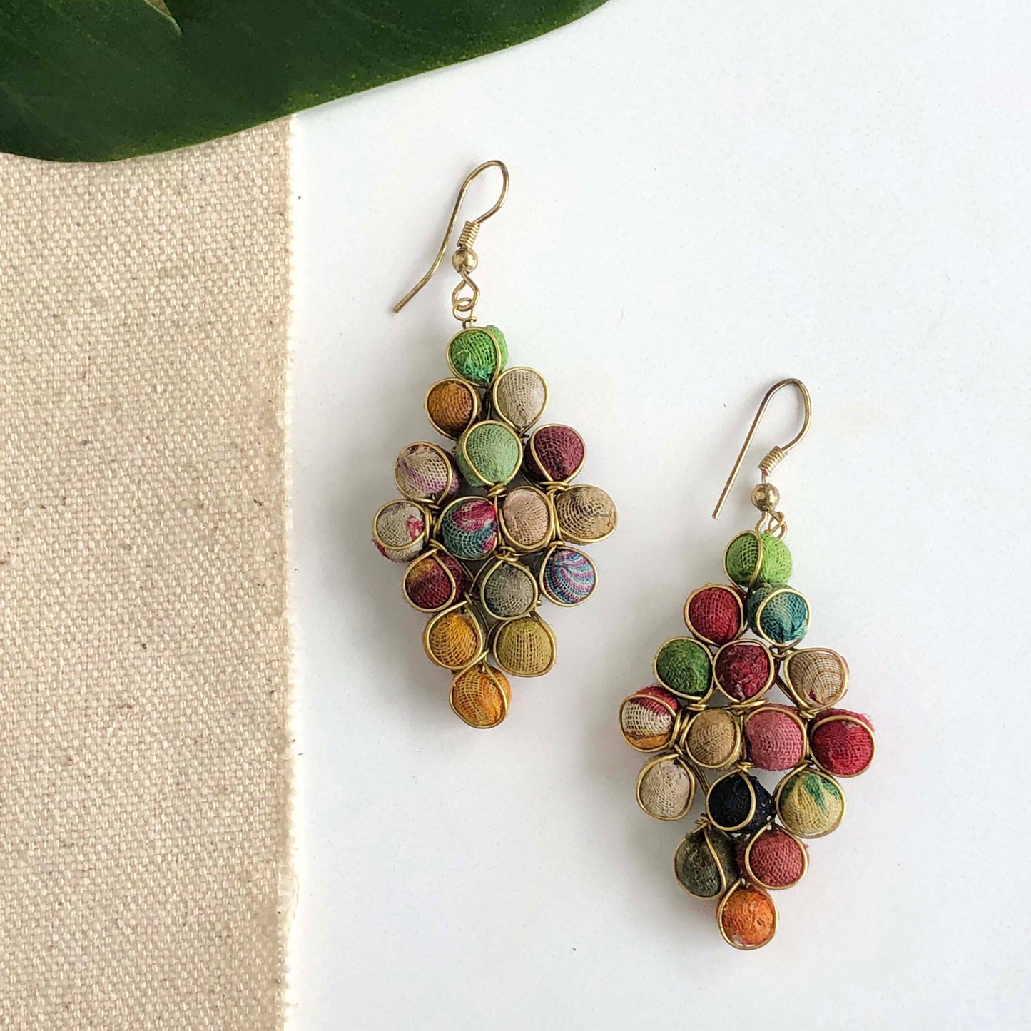 A pair of diamond-shaped earrings are formed with colorful textile-wrapped beads and gold wire.