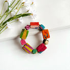 A bracelet strand of large and small square colorful textile-wrapped beads is placed against white flowers.