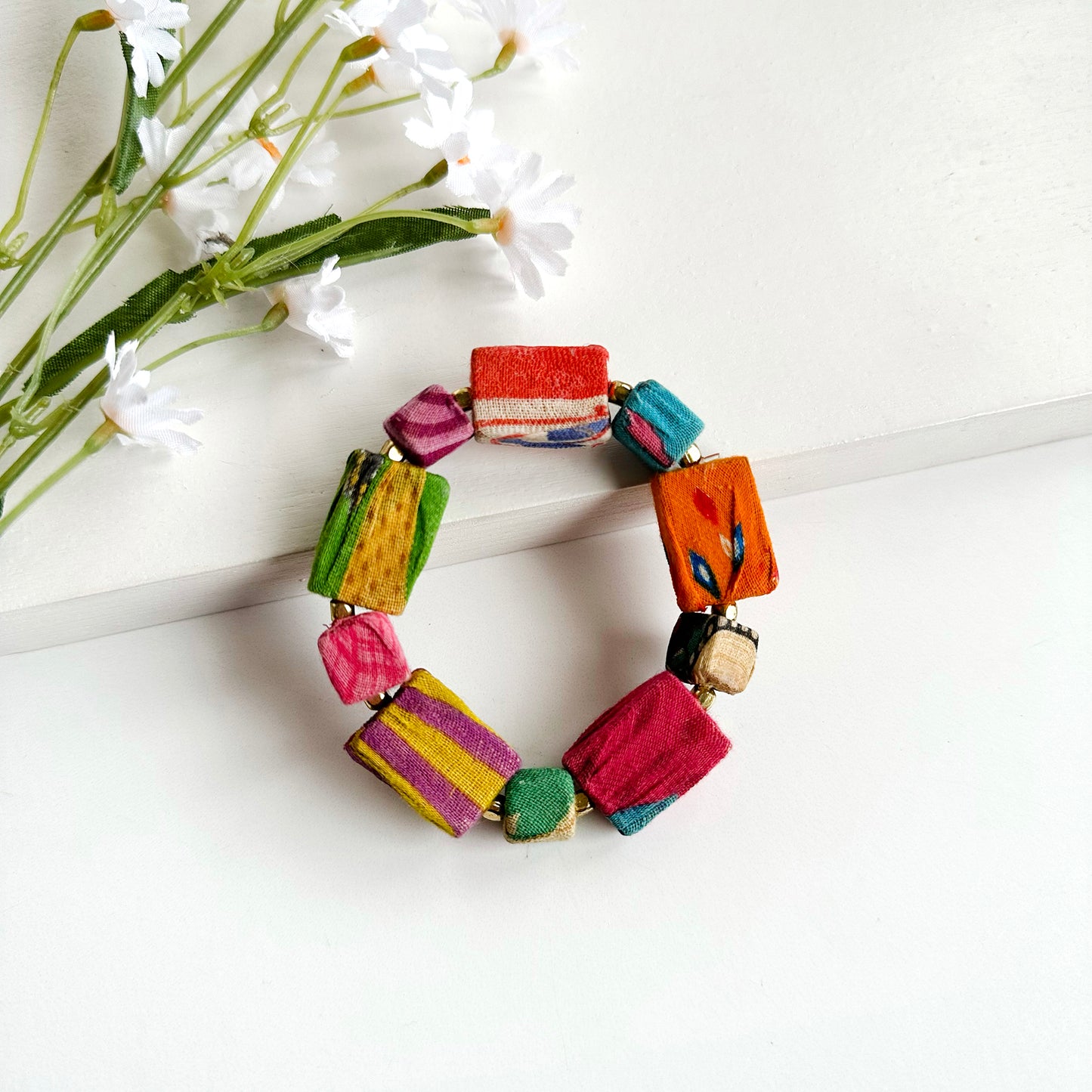 A bracelet strand of large and small square colorful textile-wrapped beads is placed against white flowers.