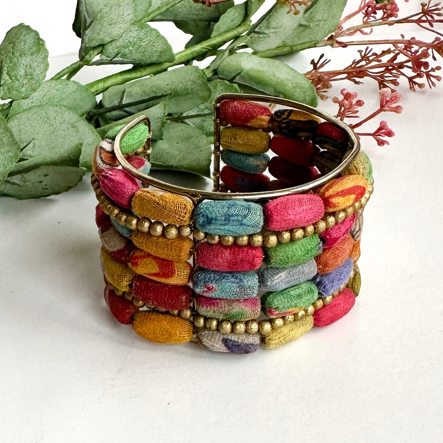 A cuff bracelet with multiple layers of beads is seen with some green leaves.
