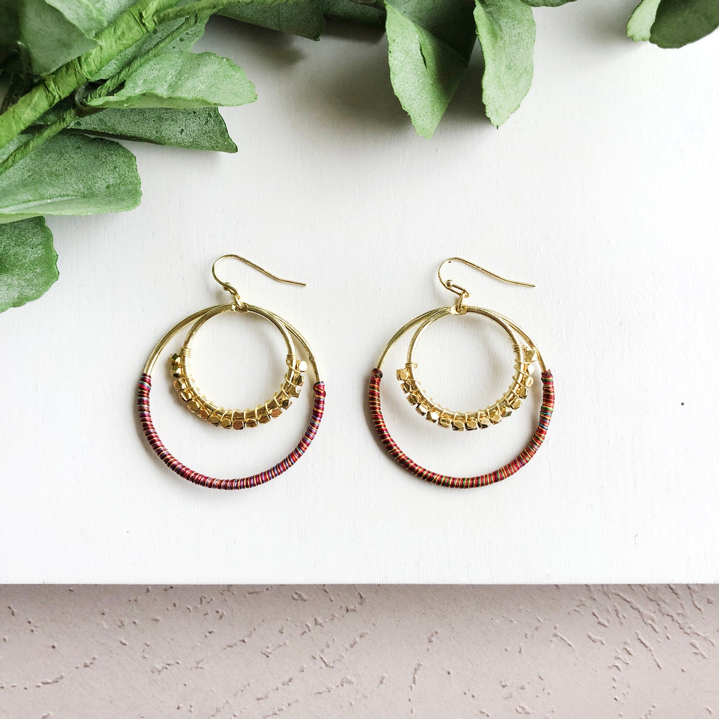 A smaller hoop embellished with faceted metallic beads layers on top of a larger rainbow thread-wrapped hoop to form a pair of earrings.