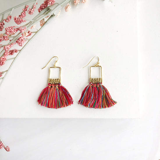 A pair of earrings is shown. The earrings are formed with a rectangular hoop, adorned at the bottom with three rows of small beads, and four long colorful tassels hang off the ends.