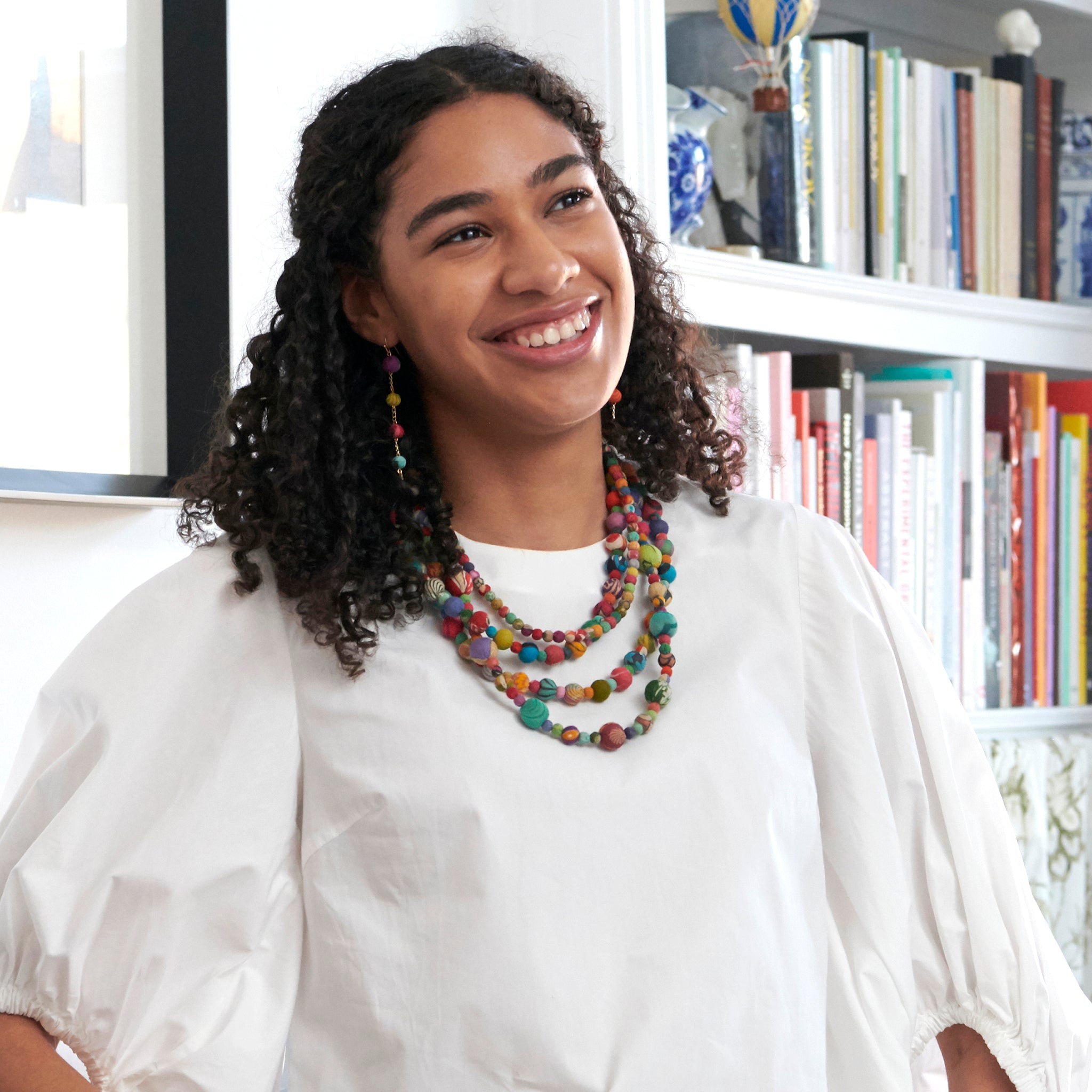 A woman smiles at us while modeling colorful beaded jewelry.