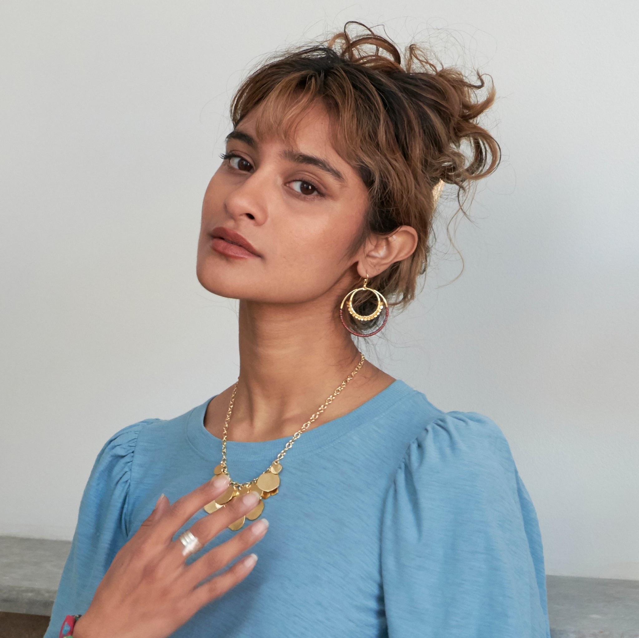 A woman looks at us while touching her necklace and modeling metallic jewelry.