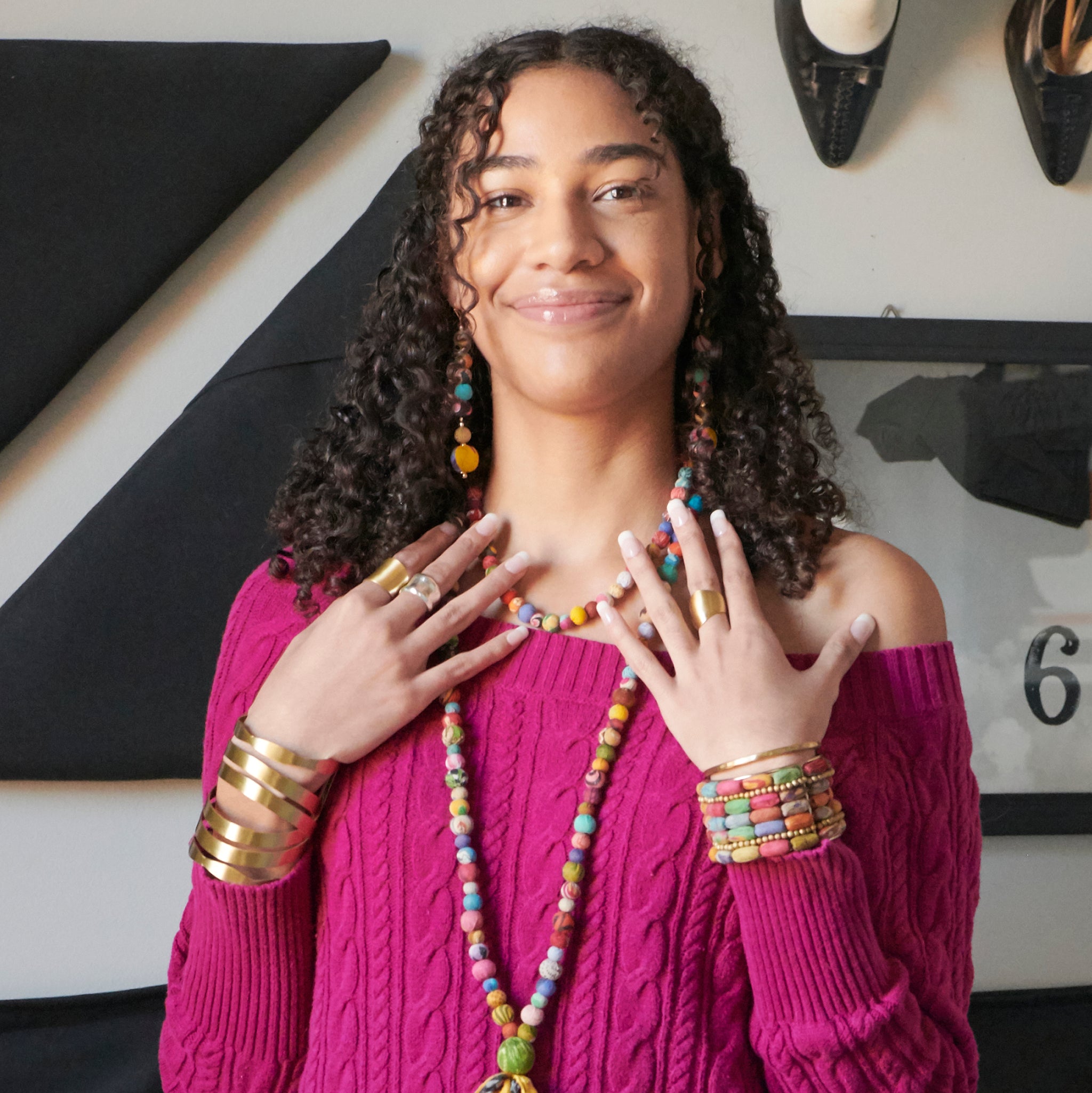 A woman places both of her hands on her collarbones and smiles at us, while adorned in various colorful jewelry.