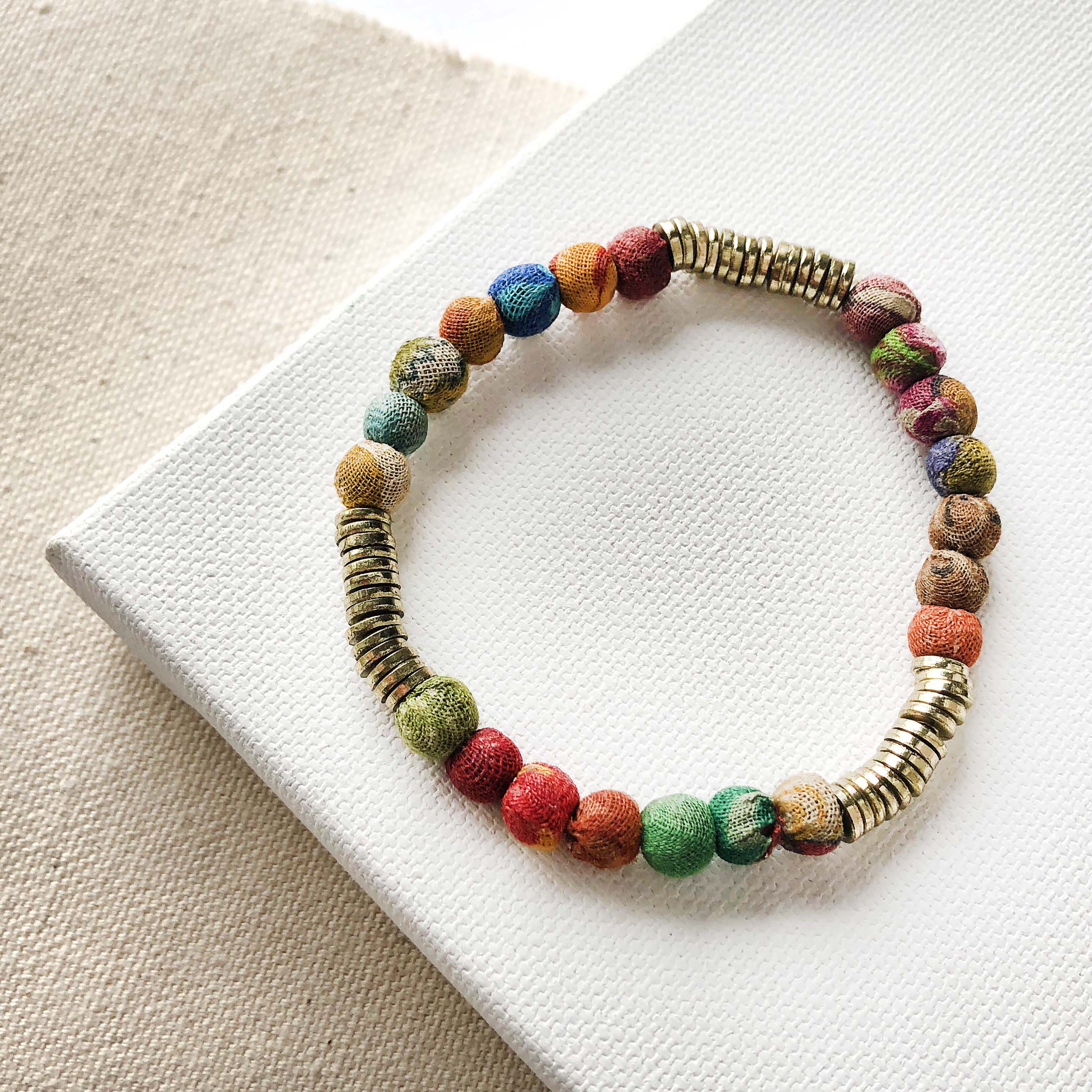A single strand bracelet of multicolor and gold beads rests on a white block against a tan background.