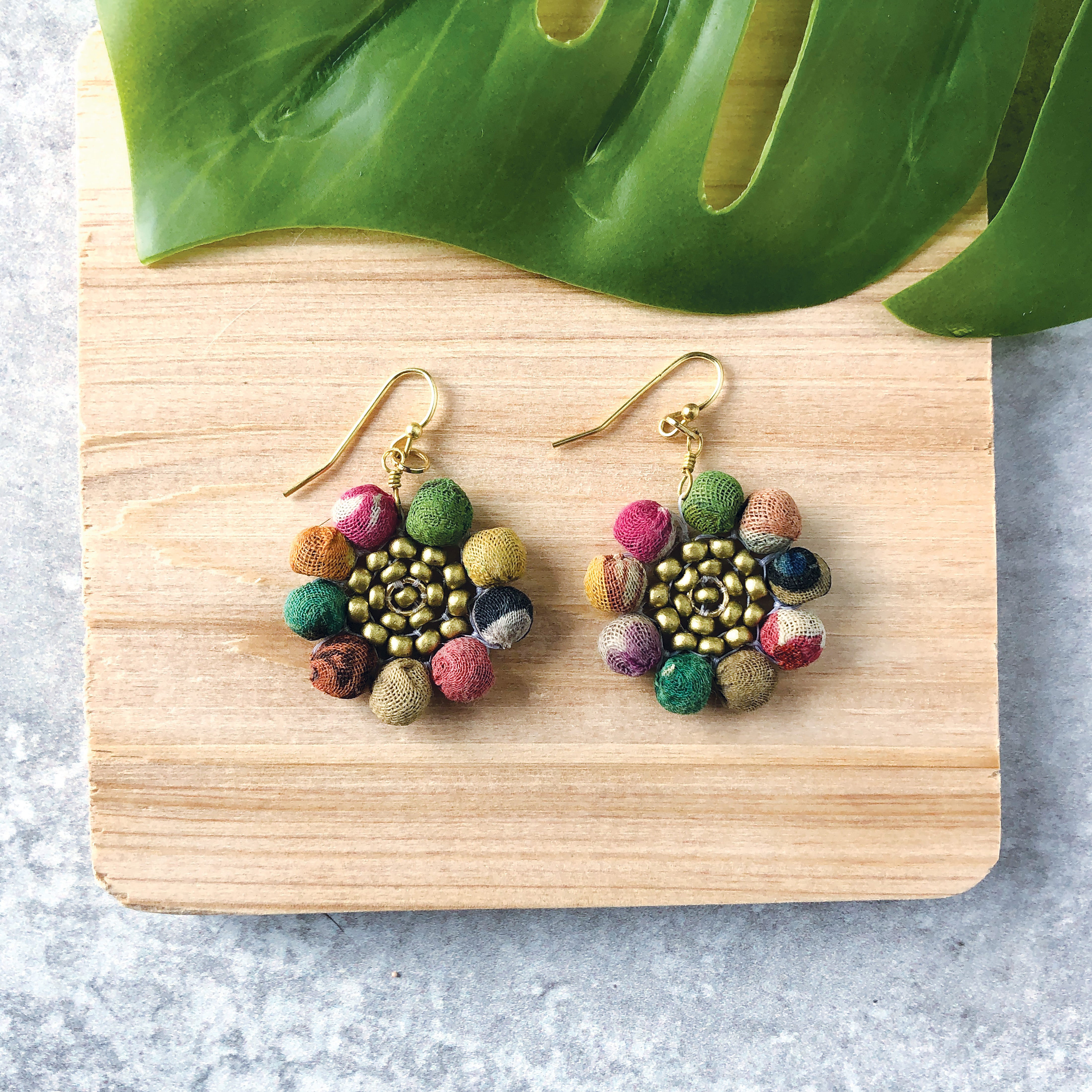 A pair of multicolor beaded earrings rests on a wooden block with a green leaf across the top.